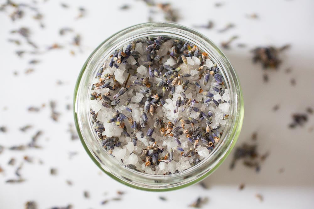 Looking over a jar of bath salt with lavender buds spread around, on a white background