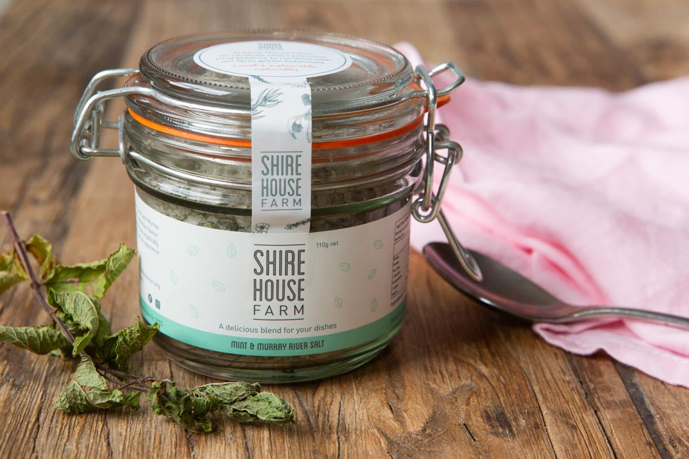 A clip top jar filled with salt, some dried mint leaves to one side and a pink cloth to the other, resting on a wood surface