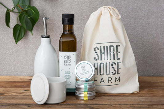 A bottle of olive oil, a ceramic pourer, three salt tins and a ceramic condiment dish, all next to a cotton pouch and on a wood surface