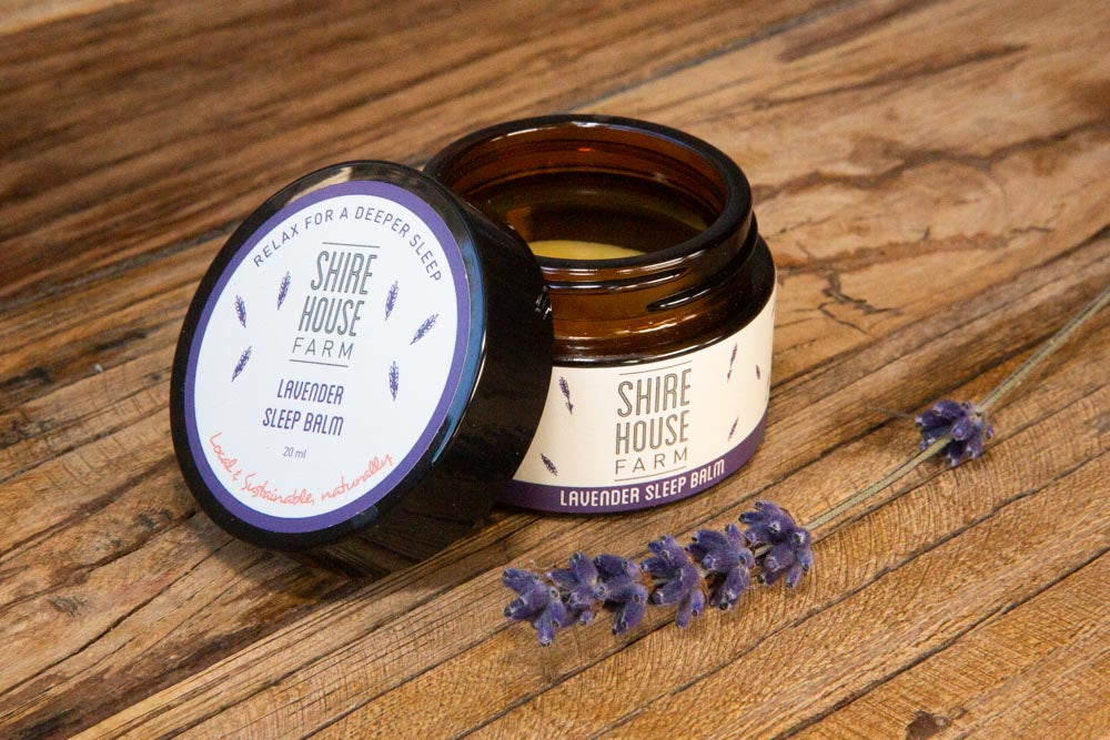 A jar of sleep balm with the lid off, a lavender sprig on the side and resting on a wood surface