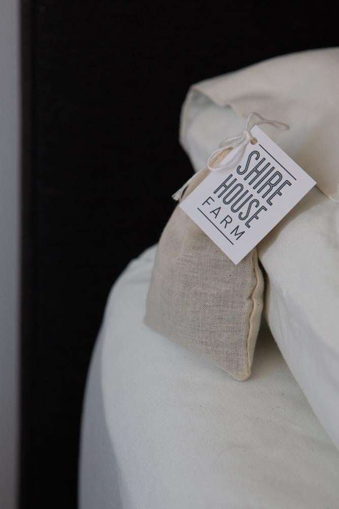 A cotton, lavender pouch with a swing tag attached, resting on a mattress, next to a pillow