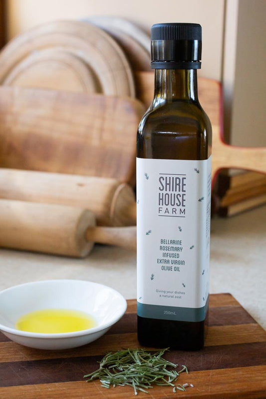Bottle of olive oil, next to a dish with olive oil and a sprinkle of rosemary leaves, on a wooden chopping board. Chopping boards in the background