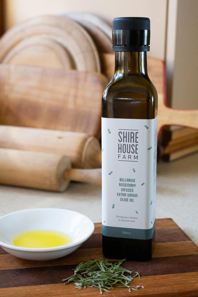 A bottle of olive oil next to a dish with oil and a sprinkle of rosemary leaves on a wooden chopping board. Chopping boards in the background