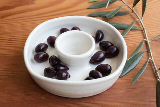 An olive dish filled with olives, an olive twig and leaves to the right, resting on a wood surface