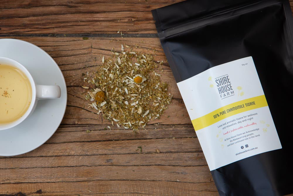 a black pouch with a white branded label, lying next to a pile of ground chamomile and two flower heads, and a cup and saucer of tea. Resting on a wood surface