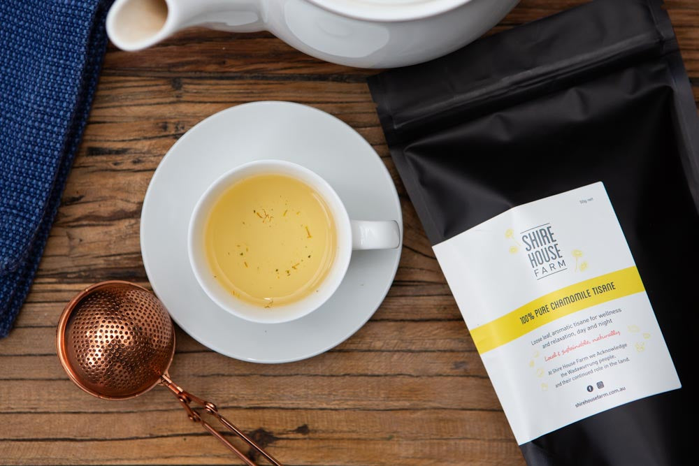 flat lay photo of black pouch with a white branded label. Lying next to a cup and saucer of tea, a teapot, rose gold tea strainer and a blue napkin. On a wood surface