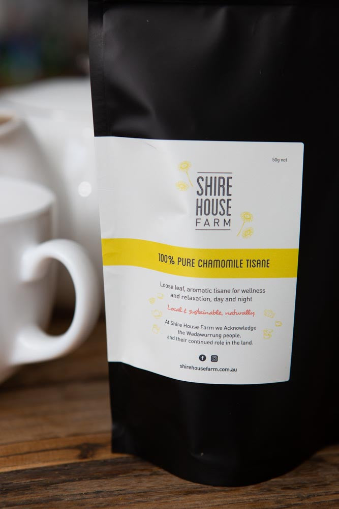 Black stand up pouch with a white branded label. Background of blurred white tea pot and mug. On a wood surface.