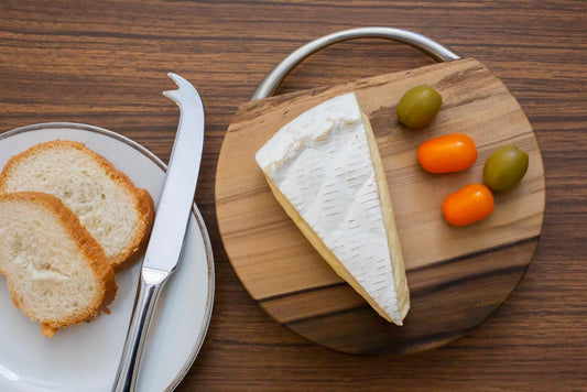 A cheese board with cheese, olives and tomatoes on it. Next to a dish with bread and a cheese knife. On a wooden bench