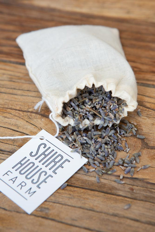 A cotton pouch open with lavender buds spilling out the top. A swing tag logo to the side, on a wood surface