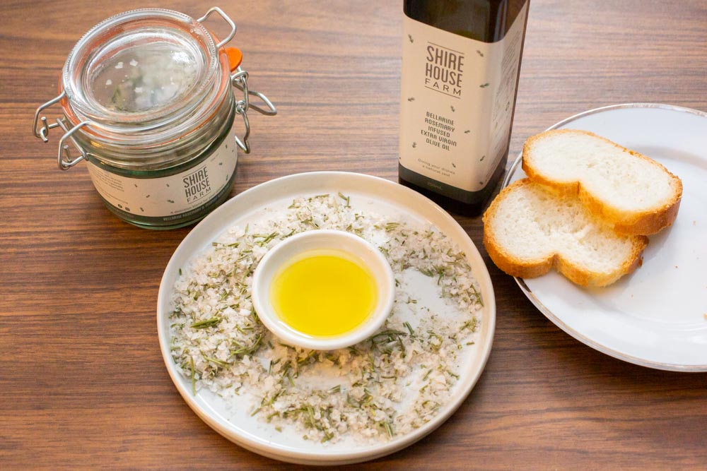 A dukkah dish filled with oil and rosemary salt. Next to a white dish with bread, and a bottle of olive oil and a clip top jar of salt. On a wooden surface