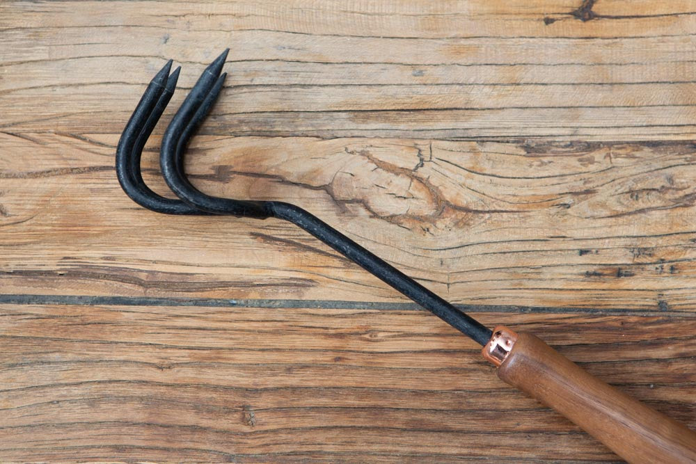 side profile of a stainless steel garden cultivator hand tool, with a wooden handle, on a wooden surface