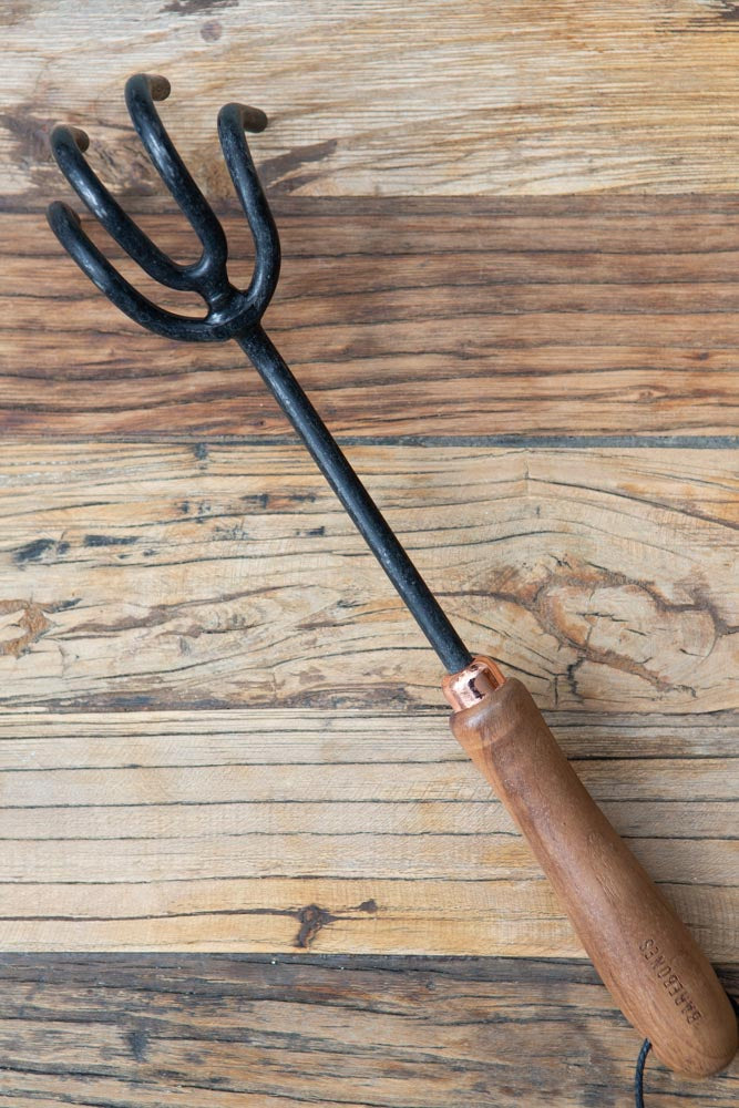 A stainless steel garden cultivator hand tool, with a wooden handle, on a wooden surface