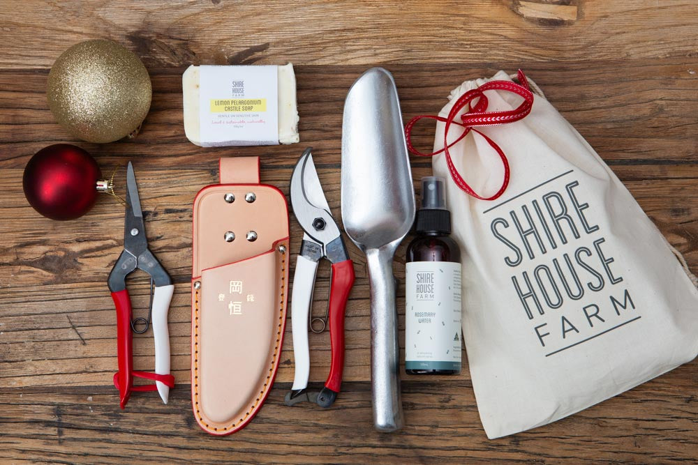 Garden tools, soap and a bottle laid flat on a wooden surface, next to a beige cotton bag. With baubles and red ribbon.