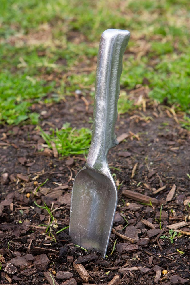 A metal garden trowel dug into the dirt, with some grass in the background