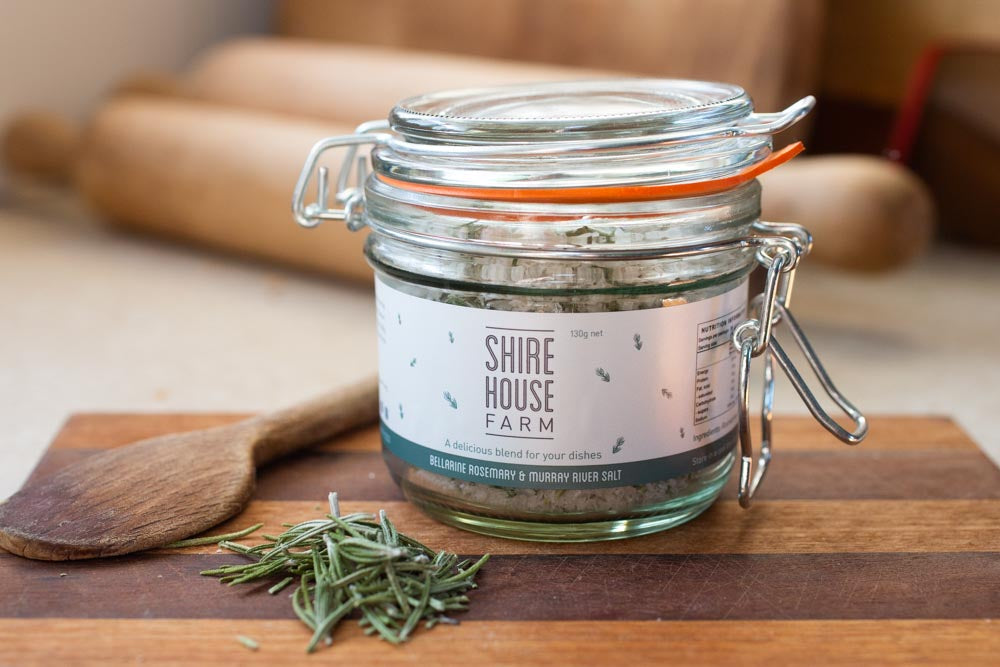 A clip top jar of salt, with a sprinkle of rosemary leaves next to it and a wooden spoon. Resting on a wooden chopping board