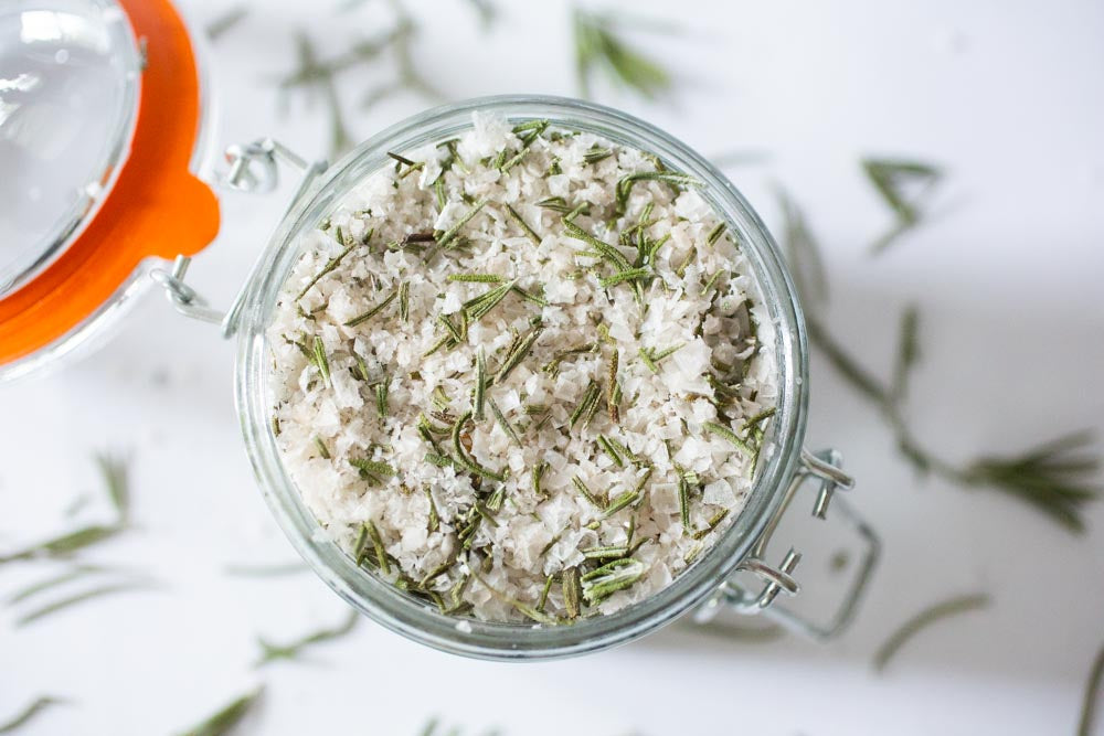 Looking over an open jar of rosemary salt with the lid off. Rosemary leaves sprinkled around the base, on a white background