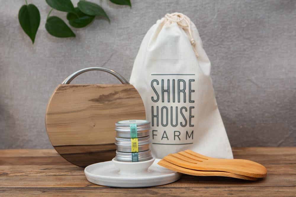 A wooden cheese board, wooden salad hands, ceramic dukkah dish and three small tins in front of a cotton back, resting on a wooden surface. Grey background with some foliage