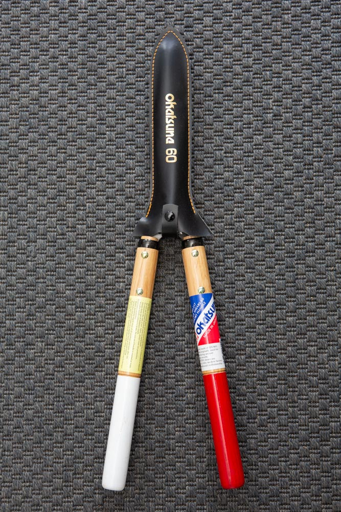 hedge shears with a black cover over the blade, red and white handles, on a dark grey textured mat