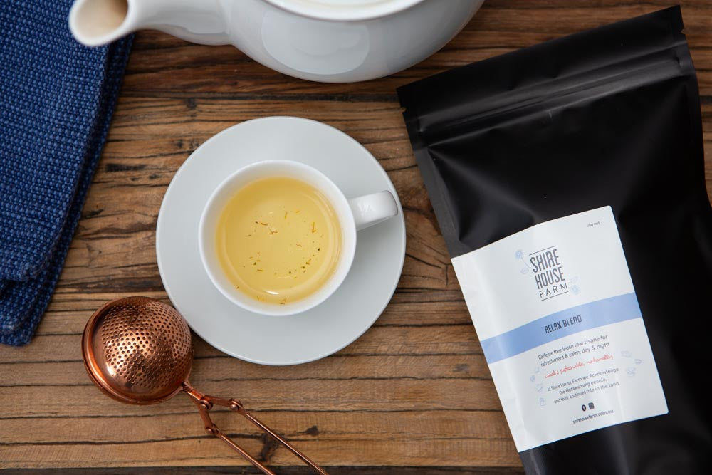 flat lay photo of a black ziplock pouch with a white branded label, next to a cup of tea on a saucer, rose gold tea strainer, white teapot and blue napkin. All on a wood surface