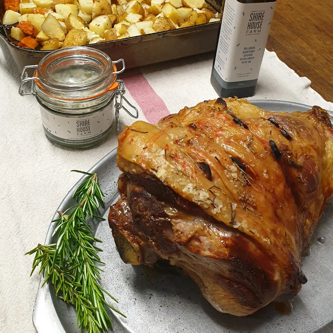 A roast lamb on a tray with a sprig of rosemary, in front of a clip top jar of salt and a bottle of olive oil, and a tray of roast potatoes
