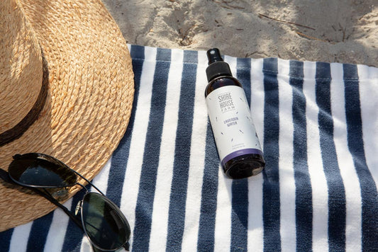 A bottle of lavender water on a blue and white striped towel, next to sunglasses and a hat, on the sand