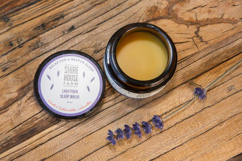 An open jar of sleep balm, with the lid to the side. A sprig of lavender resting on a wood surface