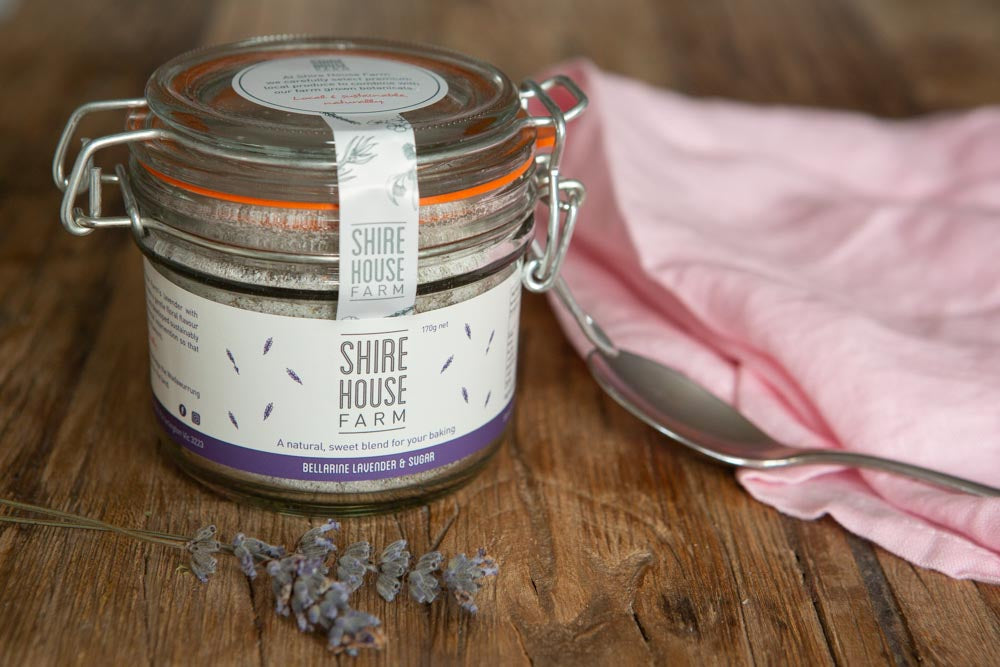 A clip top jar of lavender sugar, with a sprig of lavender to one side and a pink napkin and teaspoon to the other side. Resting on a wooden surface