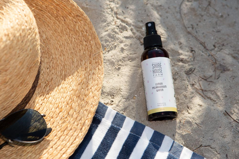 A bottle of lemon mist spray on the sand, next to a sunhat and sunglasses on a blue and white striped towel