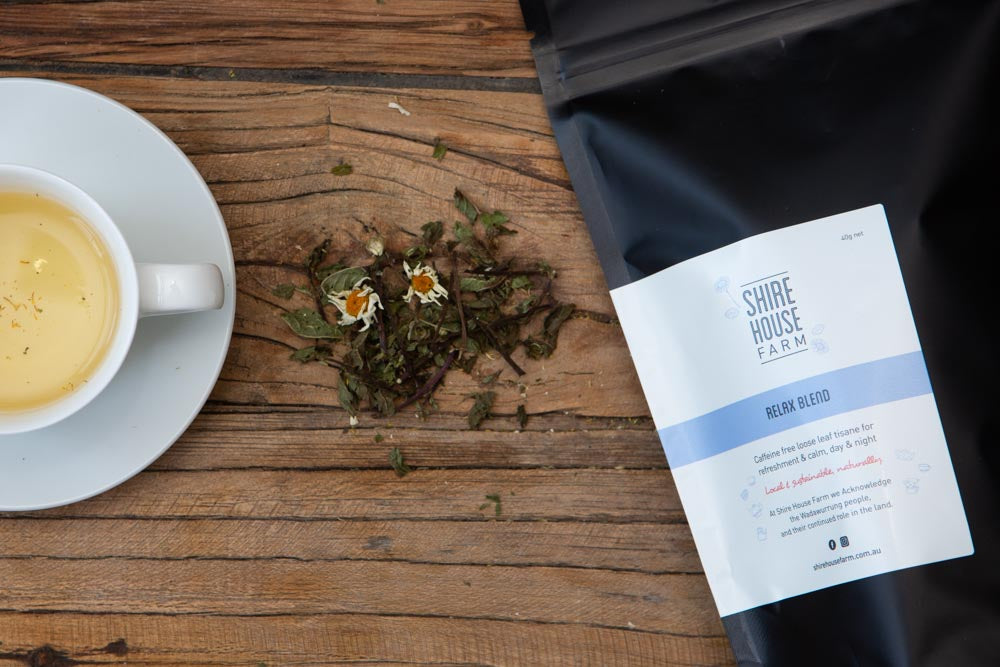 a black pouch with a white branded label, next to a pile of dried mint leaves and chamomile flowers, and a cup of tea on a saucer. All on a wood surface