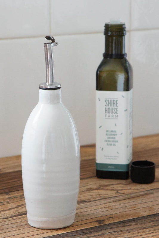 An olive oil dispenser, with a bottle of olive oil behind with the lid off. On a wood surface and white tiles behind