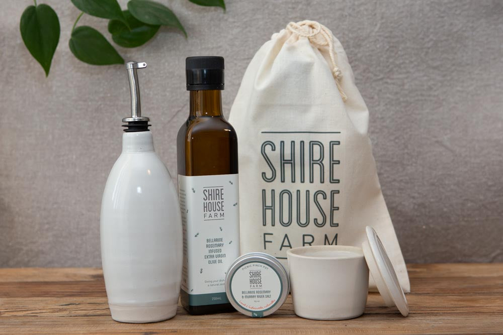 A ceramic olive oil pourer, bottle of olive oil, a ceramic dish and a tin of salt, in front of a cotton pouch, resting on a wood surface. Grey back ground with some foliage