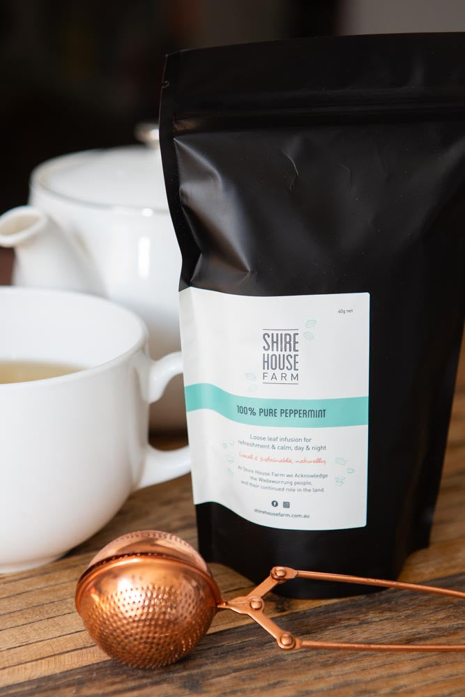 Black stand up pouch with white label, next to a white mug and teapot, on a wood surface. A rose gold tea strainer in front