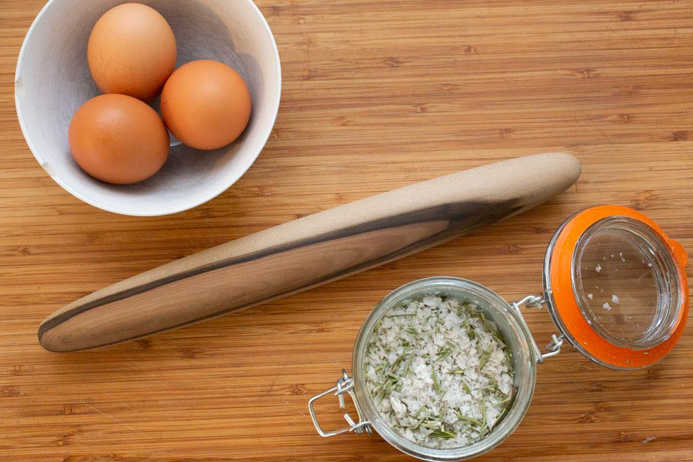 A wooden rolling pin, in between a bowl of eggs and a clip top jar of salt. Flat lay on a wood surface