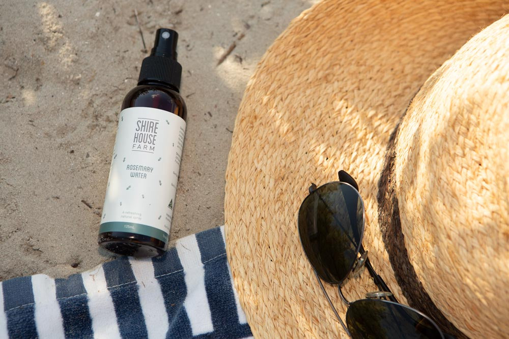 A bottle of rosemary water on the sand, next to a hat and sunglasses on a blue and white striped towel