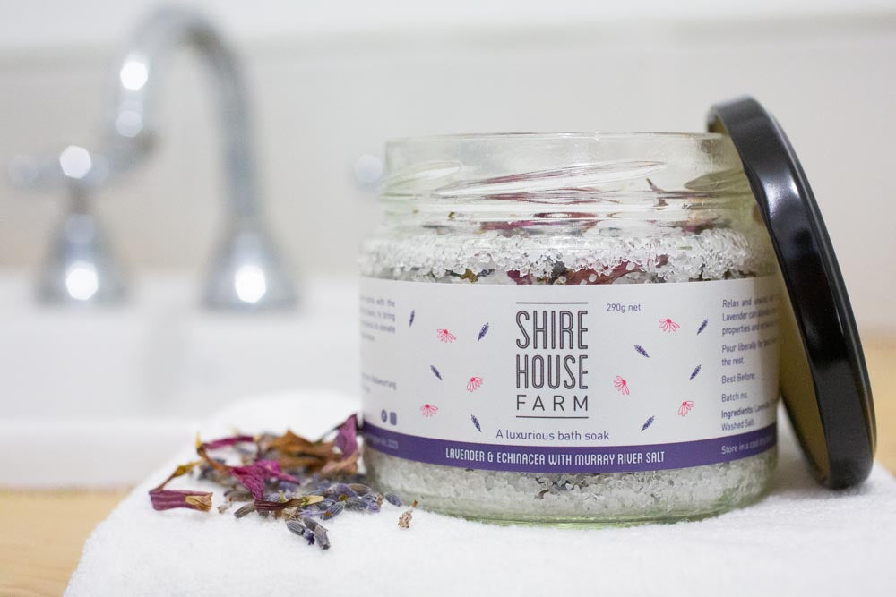 A jar of bath salt, with the lid off and flower petals on the side. Resting on a white town with a tap in the background