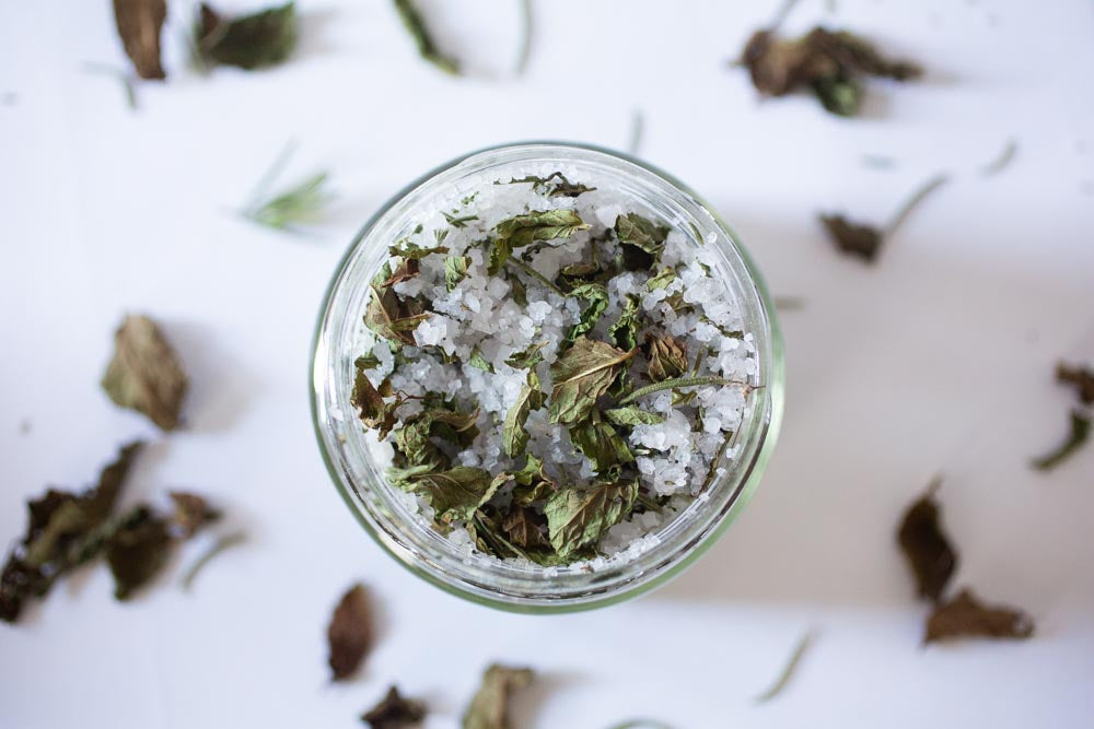 Looking over the top of an open jar of bath salt, with leaves scattered around on the white background