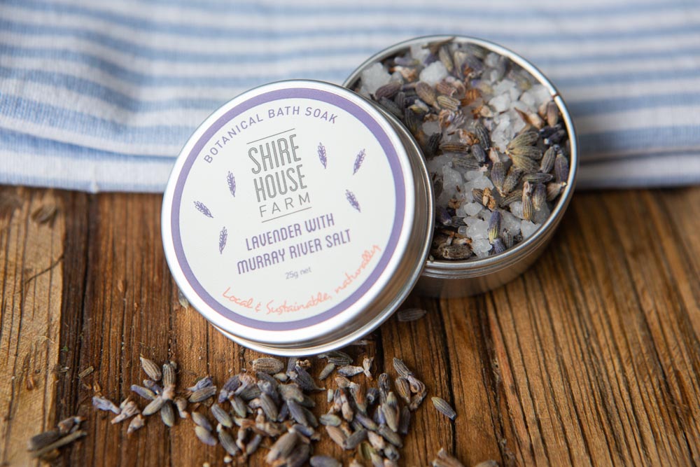 An open tin of lavender bath salt with lavender spilling out. Resting on a blue and white cloth on a wood surface