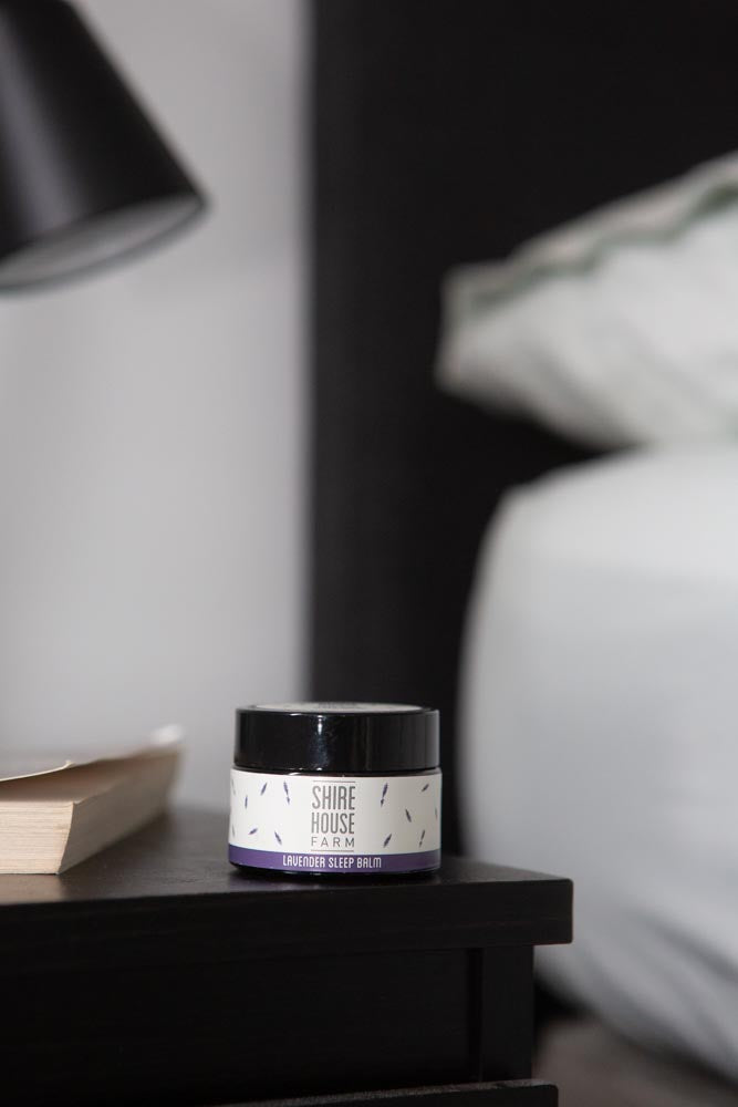 A jar of sleep balm on a black bedside table, next to a book and the corner of a bed