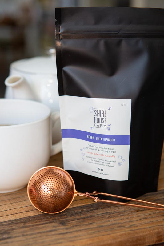 A black pouch with a white label, next to a mug and teapot, with a rose gold tea strainer in front. On a wood surface