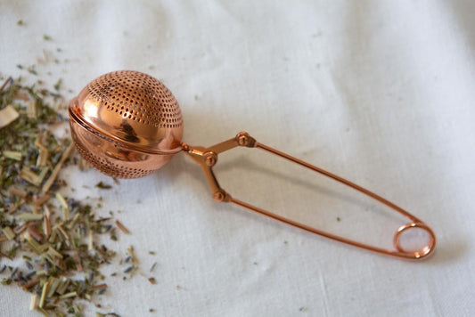 rose gold metal tea strainer with herbs laying on a white cotton surface