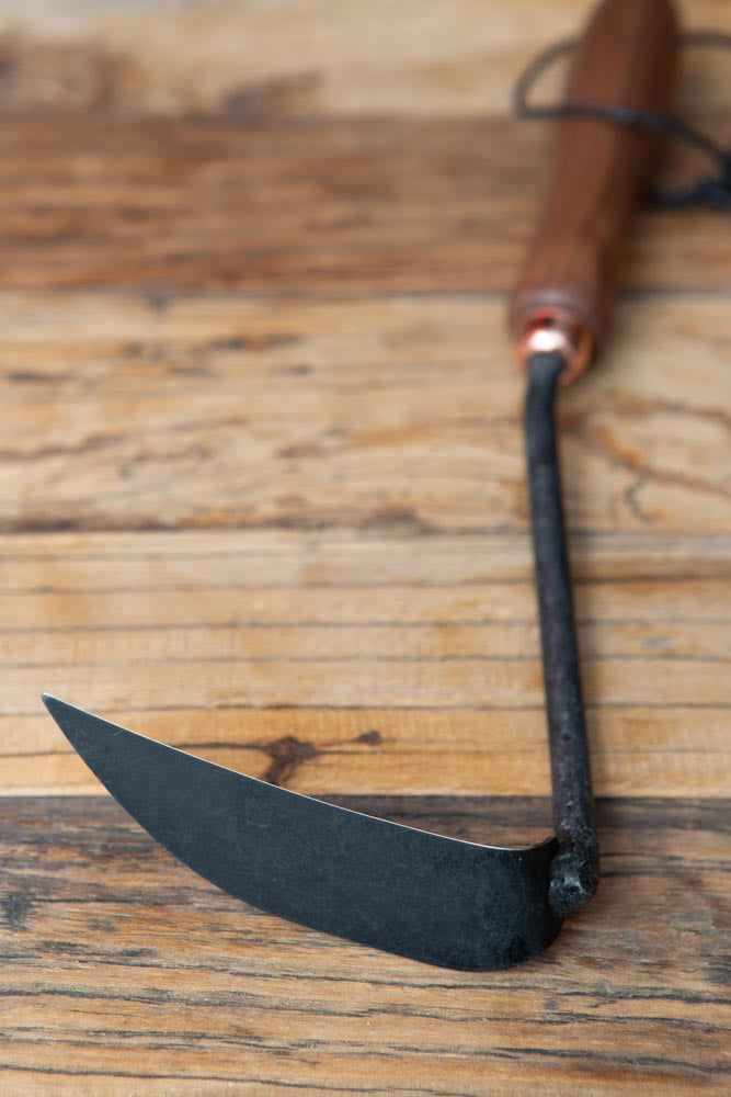 the metal part of a weeding hoe, with the wood handle blurred at the top, on a wood surface